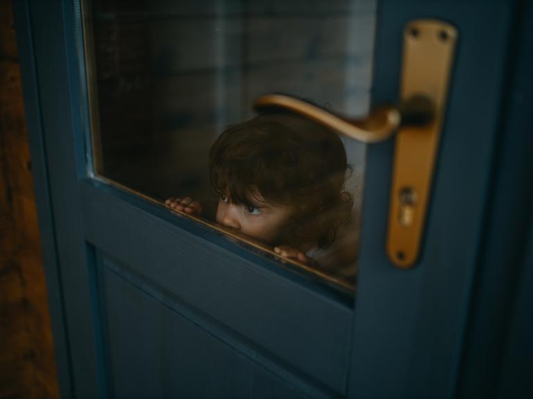 Ein Kind schaut durch die Glasscheibe einer blauen Holztür. Es reicht mit der Nase gerade bis zu der unteren Kante der Scheibe. Es sieht aus, als würde es sich verstecken.