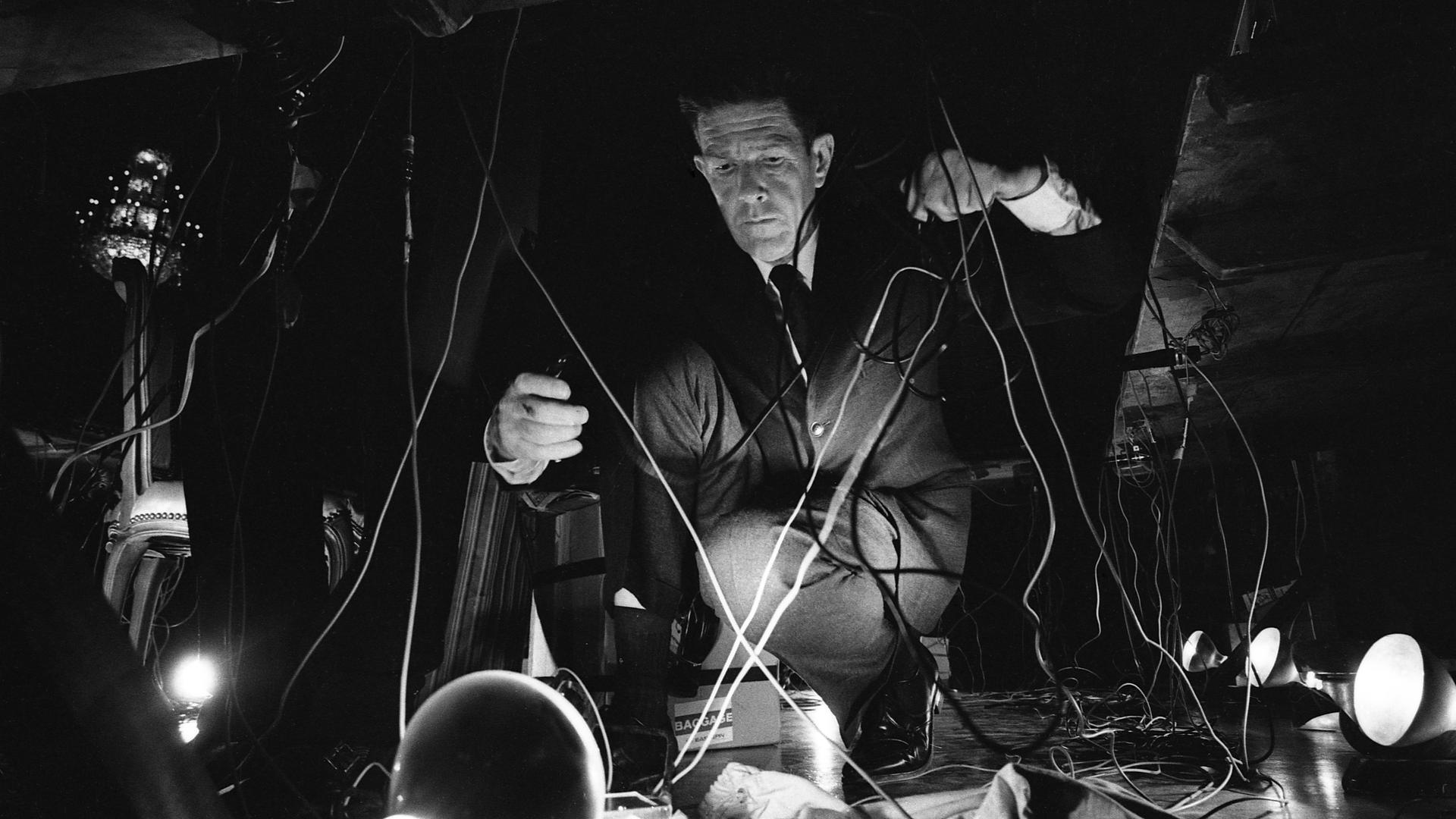 John Cage hockt während eines Konzerts auf dem Boden und hantiert mit Kabeln. John Cage hockt während eines Konzerts auf dem Boden und hantiert mit Kabeln.