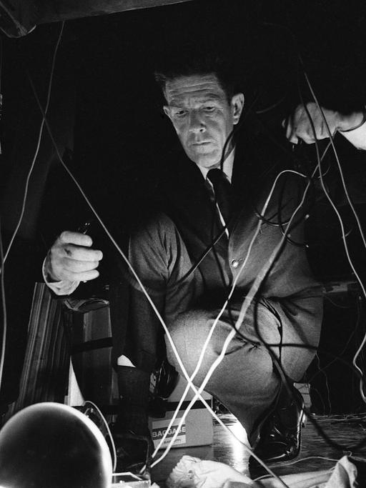 John Cage hockt während eines Konzerts auf dem Boden und hantiert mit Kabeln.