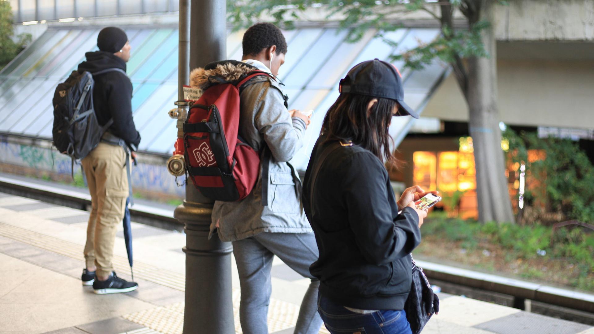 Drei Personen stehen an einem Bahnsteig. Zwei von ihnen schauen auf ihre Smartphones. Drei Personen stehen an einem Bahnsteig. Zwei von ihnen schauen auf ihre Smartphones.