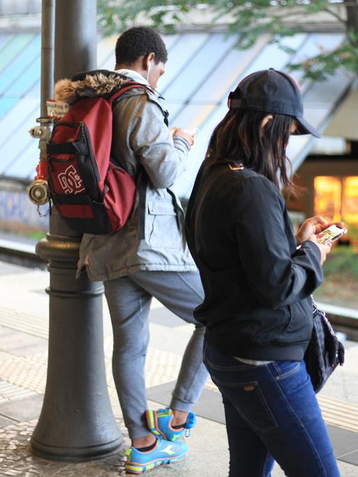 Drei Personen stehen an einem Bahnsteig. Zwei von ihnen schauen auf ihre Smartphones.