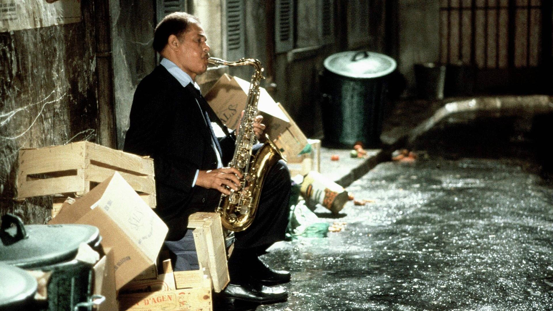 Ein Mann spielt in einem mit Kisten und Mülltonnen vollgestellten Hinterhof sitzend auf seinem Saxofon.