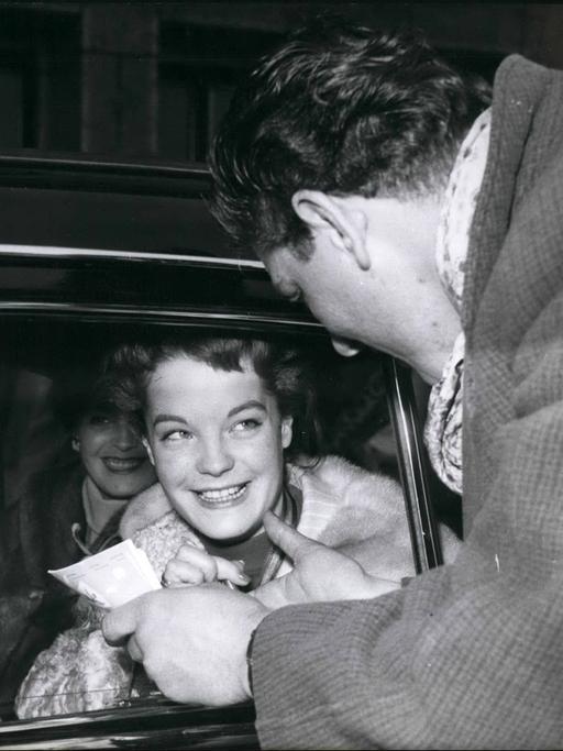 Romy Schneider. Eine junge Frau sitzt in einem Auto und wird noch einem Mann angesprochen.