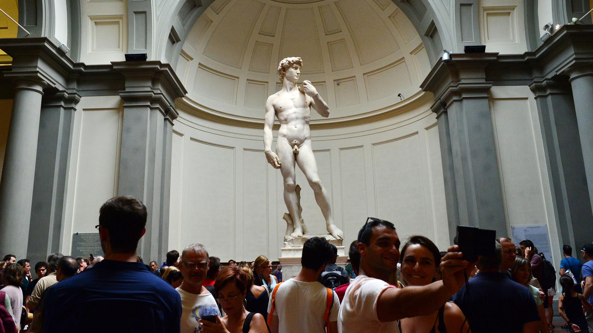 Das Foto Zeigt die David-Statue von Michelangelo in der Galleria dell Accademia in Florenz. Vor der Statue stehen viele Besucher.