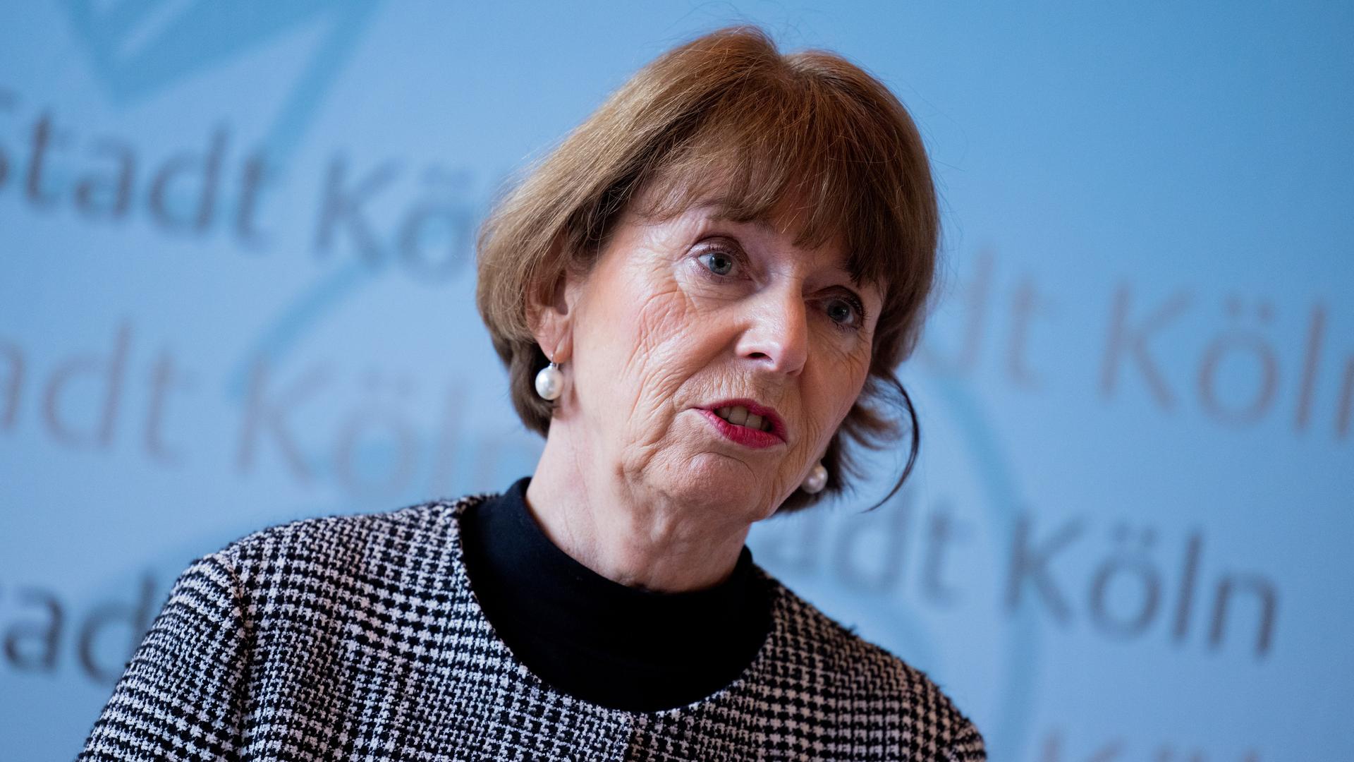 Die Kölner Oberbürgermeisterin Henriette Reker spricht mit ernster Miene vor einem hellblauen Hintergrund mit der Aufschrift "Stadt Köln".
