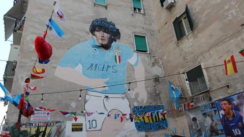 Auf einer Hauswand in Neapel prangt ein meterhohes Bild von Diego Maradona im Trikot des SSC Neapel. Der Fußballer hat schwarze, lockige Haare, das Trikot ist Blau und hat den Werbeschriftzug "Mars" auf der Brust.