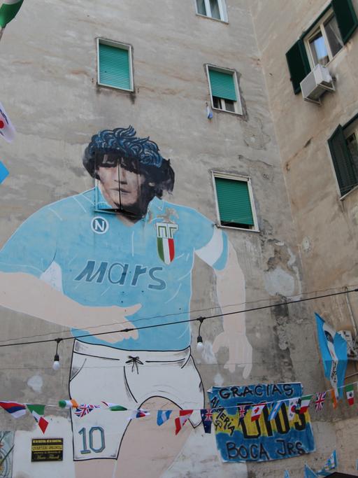 Auf einer Hauswand in Neapel prangt ein meterhohes Bild von Diego Maradona im Trikot des SSC Neapel. Der Fußballer hat schwarze, lockige Haare, das Trikot ist Blau und hat den Werbeschriftzug "Mars" auf der Brust.