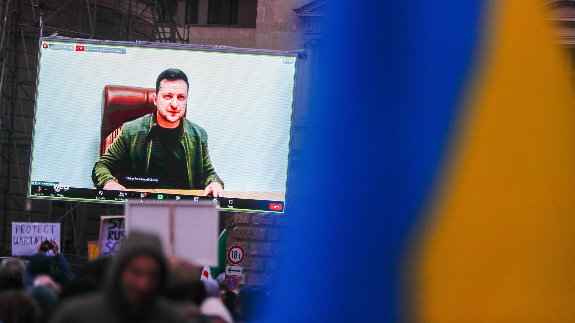Ein Bildschirm mit dem ukrainischen Präsidenten Wolodymyr Selenskyj darauf steht auf einem Platz, davor: Menschen mit Ukraine-Flaggen.