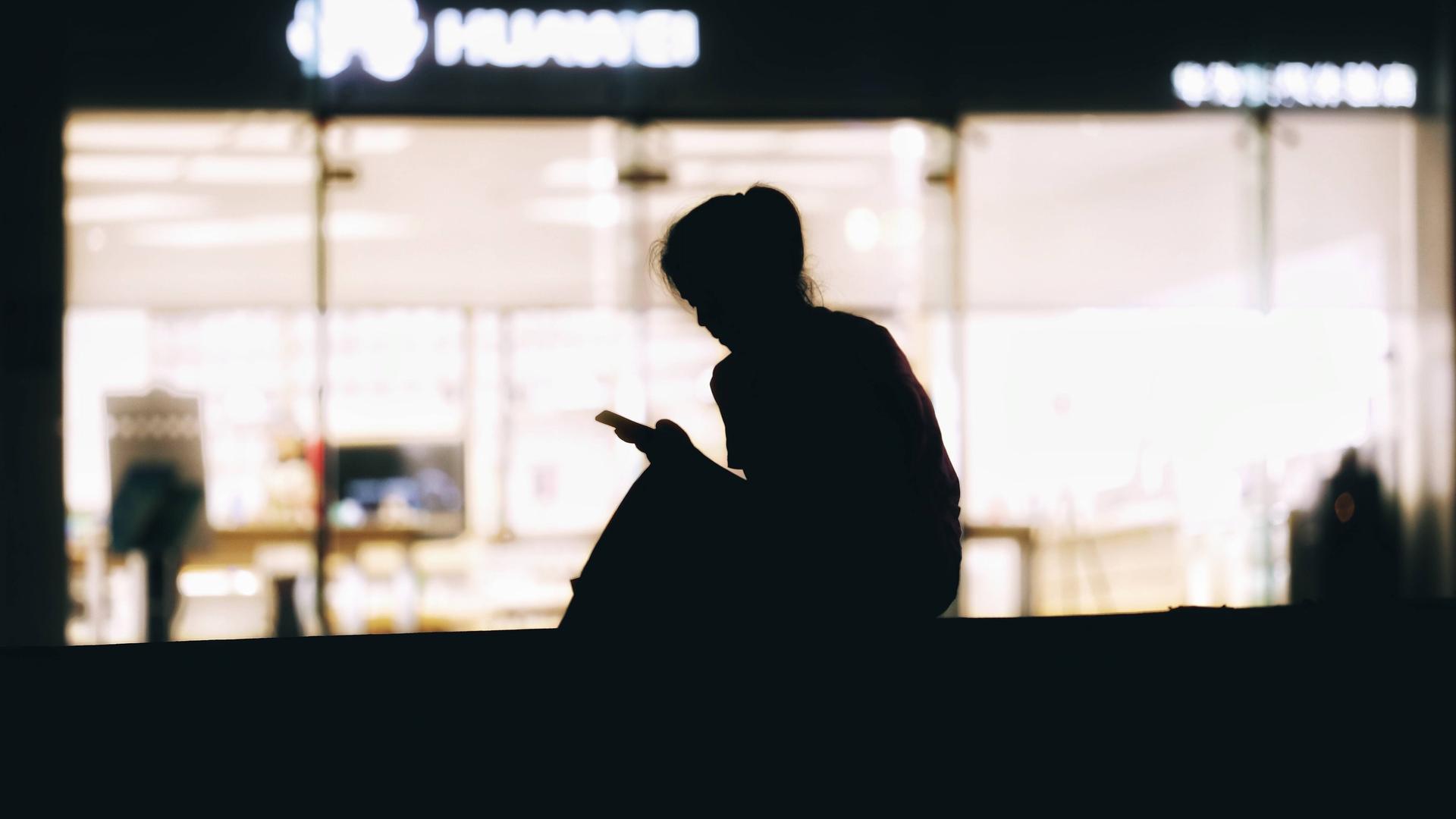 Die Silhouette einer Frau, die vor einer beleuchteten Scheibe sitzt und auf ihr Smartphone schaut. Die Silhouette einer Frau, die vor einer beleuchteten Scheibe sitzt und auf ihr Smartphone schaut.