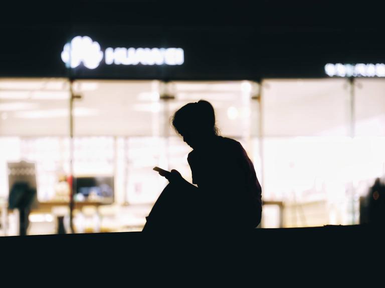Die Silhouette einer Frau, die vor einer beleuchteten Scheibe sitzt und auf ihr Smartphone schaut. 