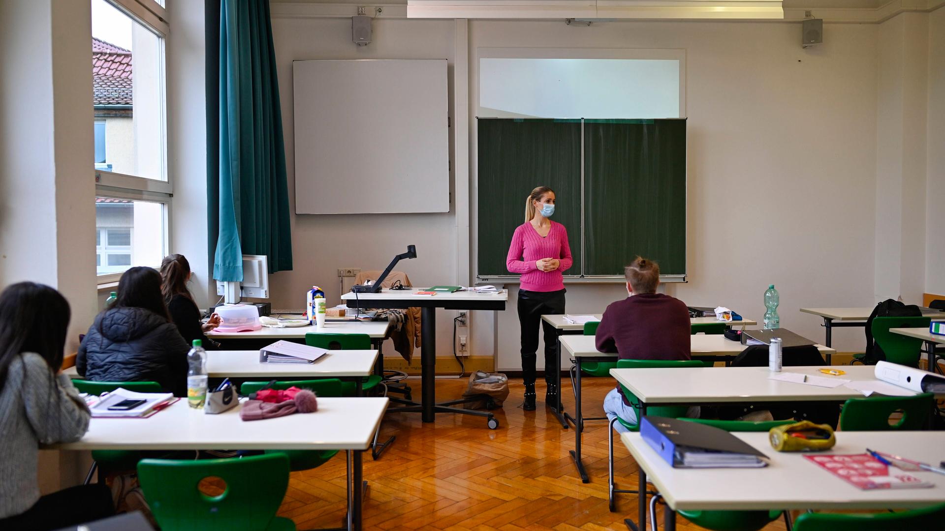 Eine Lehrerin mit Mundschutz steht an der Tafel. Von den Schülern ist jeder Tisch nur einzeln besetzt. Eine Lehrerin mit Mundschutz steht an der Tafel. Von den Schülern ist jeder Tisch nur einzeln besetzt.