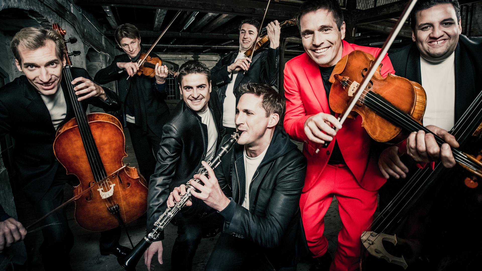 Sieben Musiker spielen vergnügt ihre Instrumente. Einer der Männer trägt einen leuchtend roten Anzug, der Rest trägt schwarz-weiße Kleidung. Sieben Musiker spielen vergnügt ihre Instrumente. Einer der Männer trägt einen leuchtend roten Anzug, der Rest trägt schwarz-weiße Kleidung.