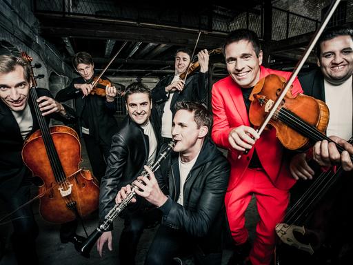 Sieben Musiker spielen vergnügt ihre Instrumente. Einer der Männer trägt einen leuchtend roten Anzug, der Rest trägt schwarz-weiße Kleidung.