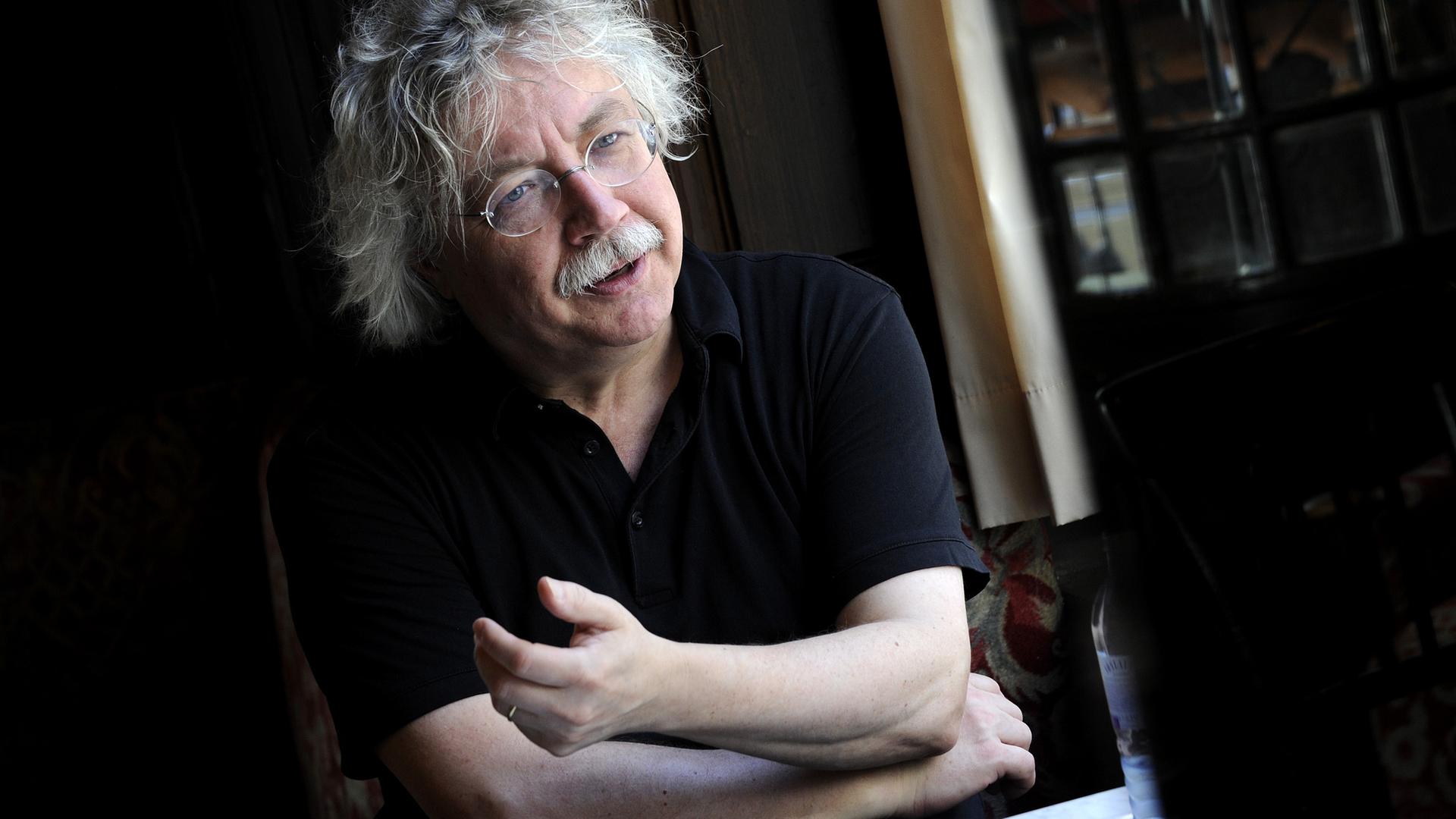 Der Schriftsteller Karl-Markus Gauß, in einem schwarzen Polohemd, mit weißem lockigem Haar und weißem Schnurrbart, scheint mitten im Gespräch zu sein, gerade hebt er die linke Hand ein wenig vom Tisch, als wollte er ein Argument unterstreichen. Der Schriftsteller Karl-Markus Gauß, in einem schwarzen Polohemd, mit weißem lockigem Haar und weißem Schnurrbart, scheint mitten im Gespräch zu sein, gerade hebt er die linke Hand ein wenig vom Tisch, als wollte er ein Argument unterstreichen.