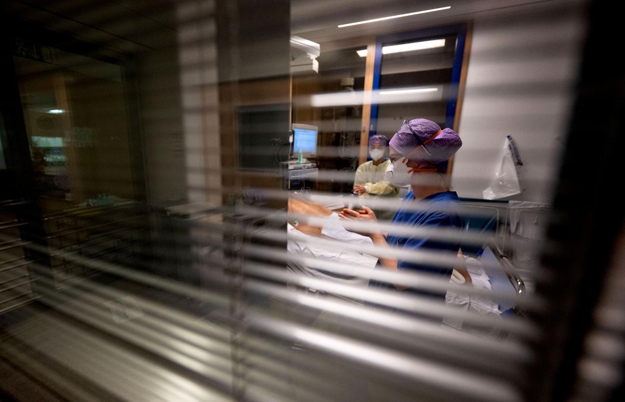 Durch ein Fenster mit halb geschlossenen Jalousien werden Krankenschwestern beobachtet, die sich auf der Intensivstation um einen Komapatienten kümmern. Durch ein Fenster mit halb geschlossenen Jalousien werden Krankenschwestern beobachtet, die sich auf der Intensivstation um einen Komapatienten kümmern.