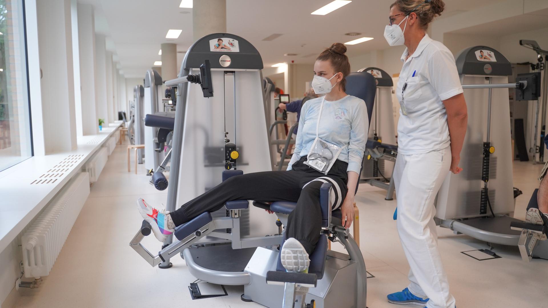 Eine Long-Covid-Patientin sitzt auf einer Beinpresse. Neben ihr steht eine Sporttherapeutin. Eine Long-Covid-Patientin sitzt auf einer Beinpresse. Neben ihr steht eine Sporttherapeutin.
