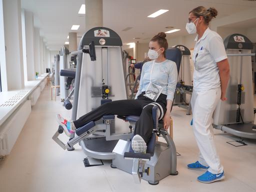Eine Long-Covid-Patientin sitzt auf einer Beinpresse. Neben ihr steht eine Sporttherapeutin.