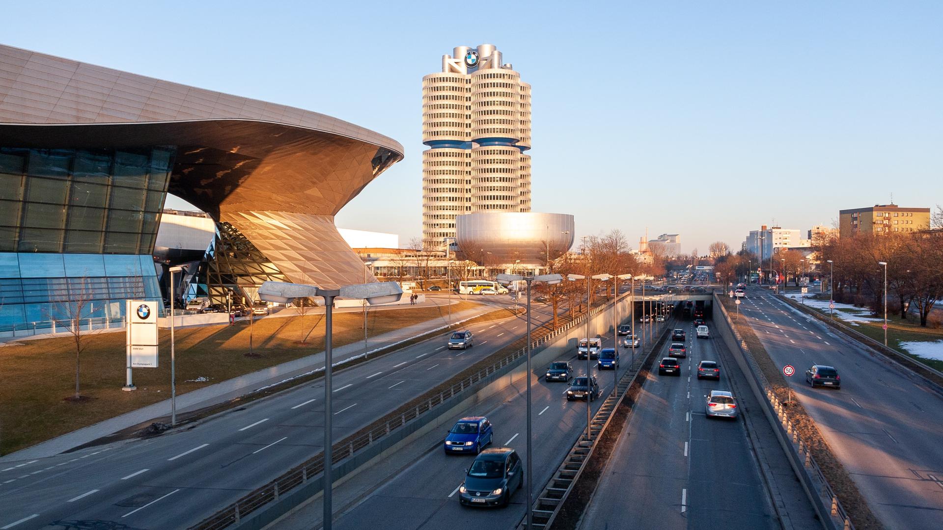 Autos befahren eine Straße, im Hintergrund ist das Hauptgebäude von BMW zu sehen, dessen Architektur an einen Vierzylindermotor angelehnt ist.