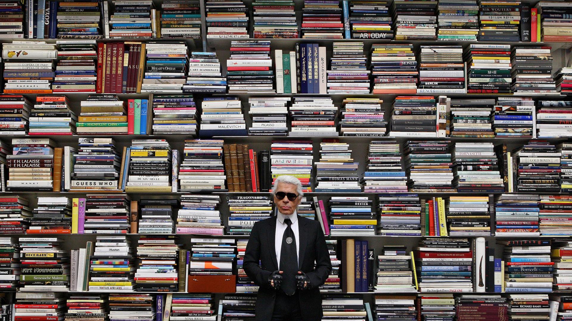 Ein Mann in schwarzer Kleidung und mit Sonnenbrille steht vor einem riesigen Regal mit hunderten von großformatigen Büchern. Ein Mann in schwarzer Kleidung und mit Sonnenbrille steht vor einem riesigen Regal mit hunderten von großformatigen Büchern.