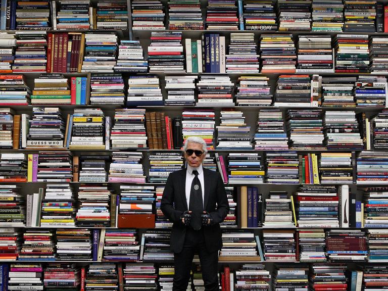 Ein Mann in schwarzer Kleidung und mit Sonnenbrille steht vor einem riesigen Regal mit hunderten von großformatigen Büchern.