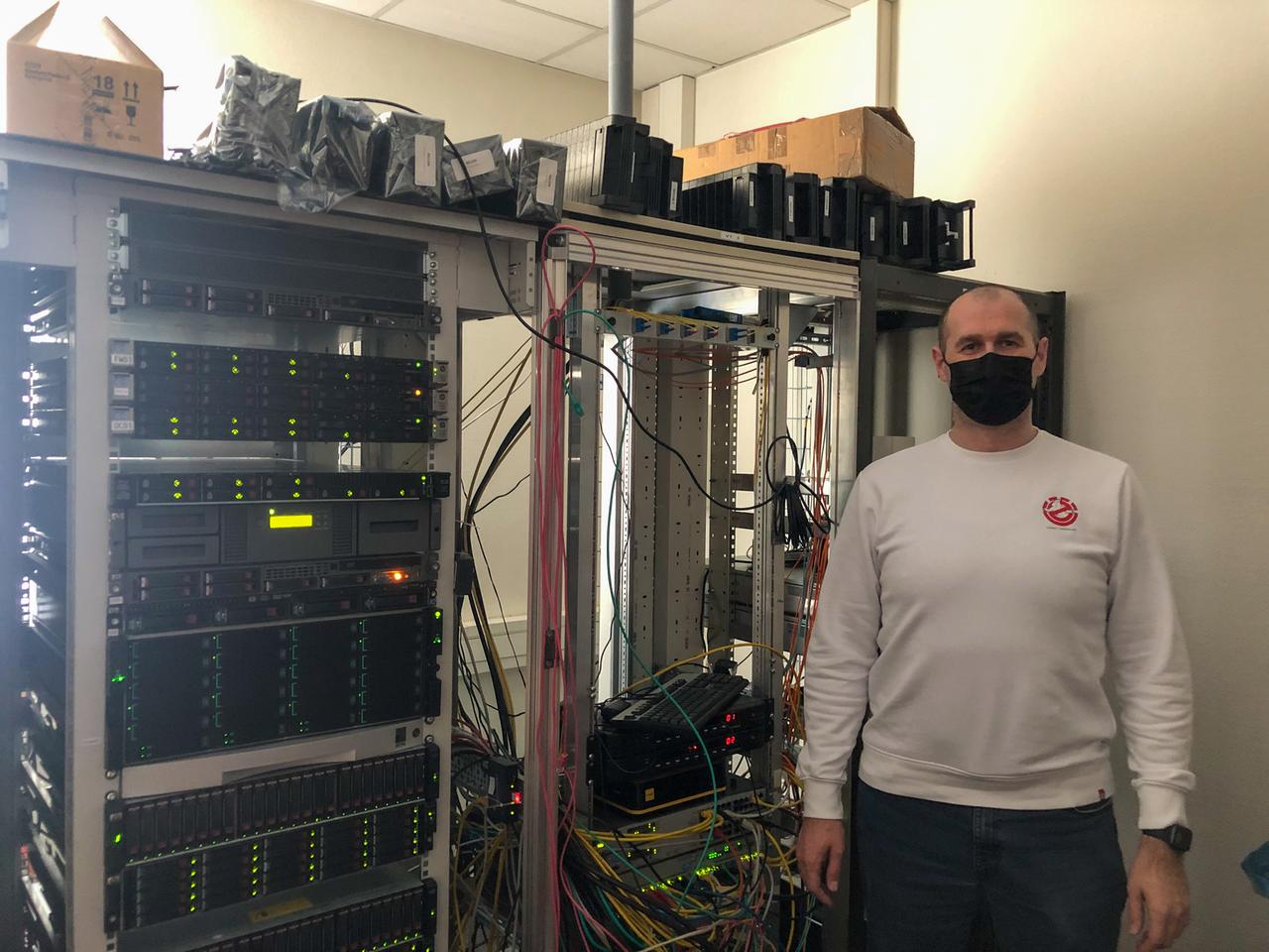 Ein Mann mit Maske steht vor einem großen Serverschrank und schaut in die Kamera