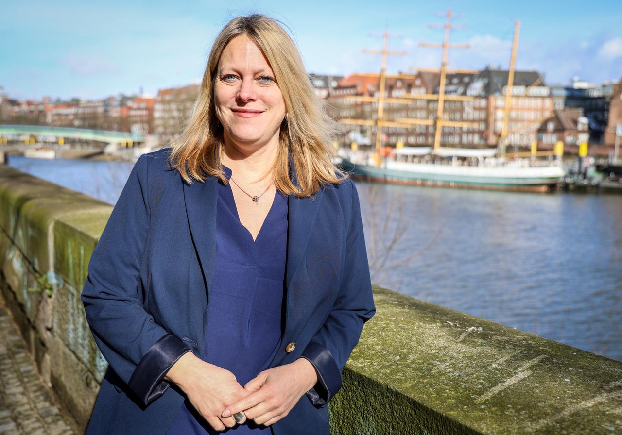 Eine junge Frau mit langen blonden Haaren steht im Freien. Im Hintergrund ist Wasser und Bremen zu sehen. 