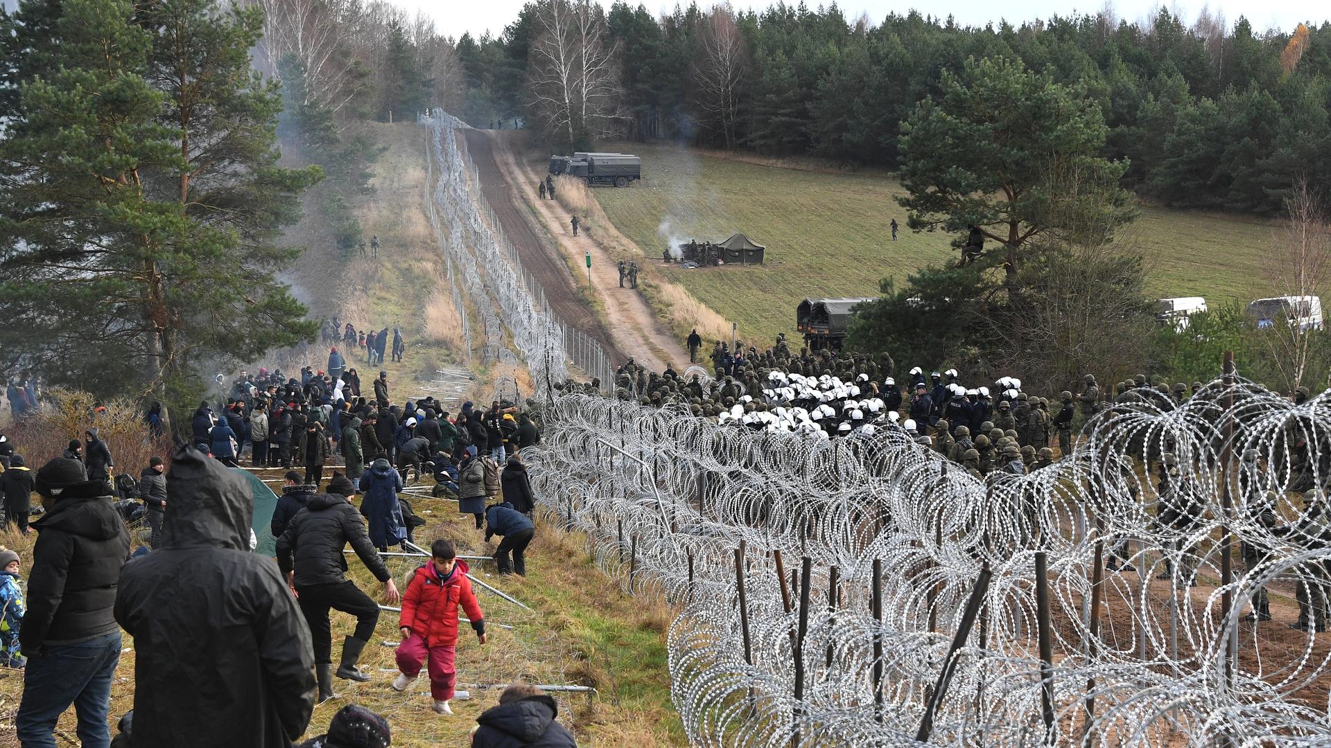 Dutzende Flüchtlinge stehen an der belarussisch-polnischen Grenze, getrennt durch einen Stacheldrahtzaun, dutzenden Polizeikräften gegenüber. 