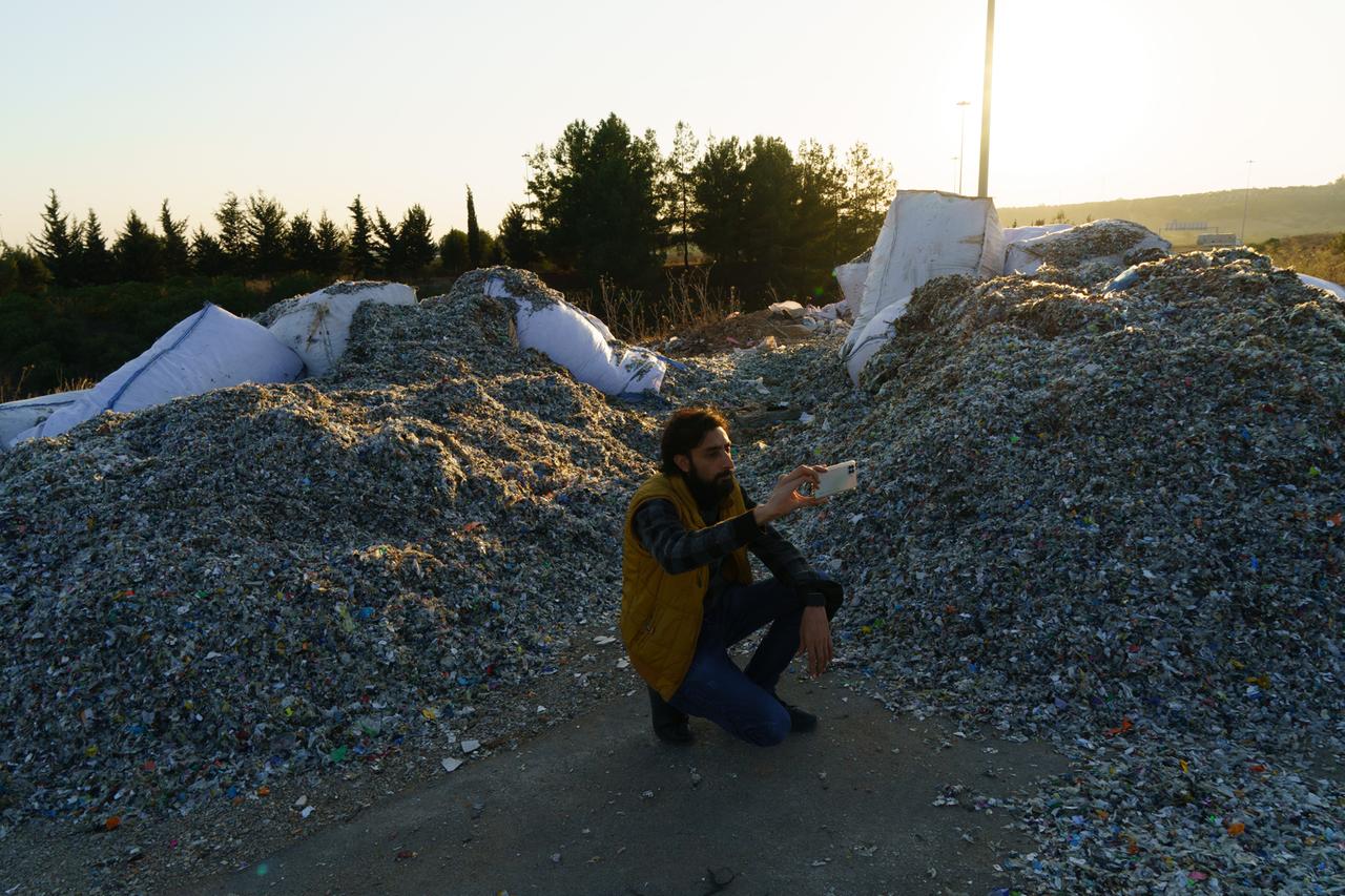 Ein Mann mit Bart und Jeans kniet zwischen zwei Abfallbergen und macht Fotos mit seinem Smartphone. Ein Mann mit Bart und Jeans kniet zwischen zwei Abfallbergen und macht Fotos mit seinem Smartphone.