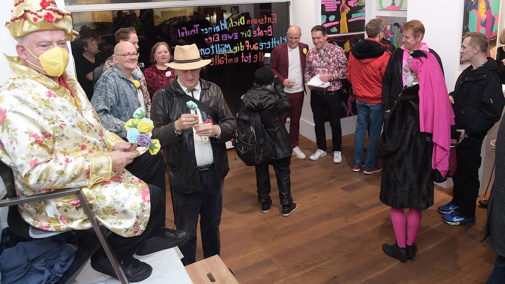 Rosa von Praunheim thront bei der Vernissage seiner Ausstellung in Berlin auf einem Stuhl, der auf einem Tisch steht. Er hat einen Spitzhut auf und ein Gewand in passenden Farben, er trägt eine Maske zum Schutz vor einer Corona-Ansteckung. Rosa von Praunheim thront bei der Vernissage seiner Ausstellung in Berlin auf einem Stuhl, der auf einem Tisch steht. Er hat einen Spitzhut auf und ein Gewand in passenden Farben, er trägt eine Maske zum Schutz vor einer Corona-Ansteckung.
