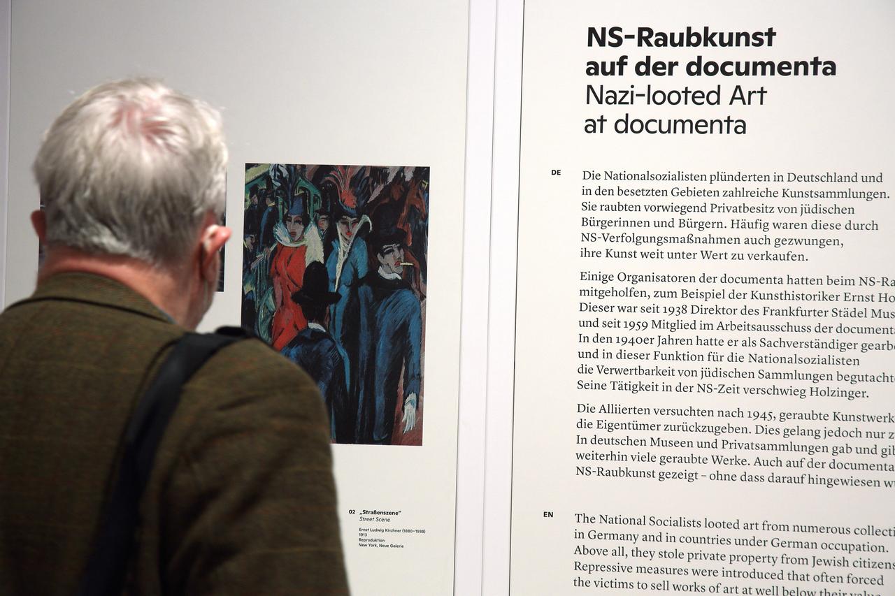 Eine Person, von hinten zu sehen, betrachtet eine Ausstellungswand mit der Überschrift 2NS-Raubkunst auf der documenta". Daneben ist ein Bild von Ernst Ludwig Kirchner zu sehen.