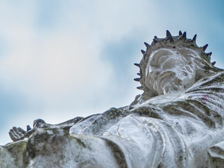 Blick nach oben zu einem steinernen Cruzifix. Jesus trägt eine Dornenkrone.