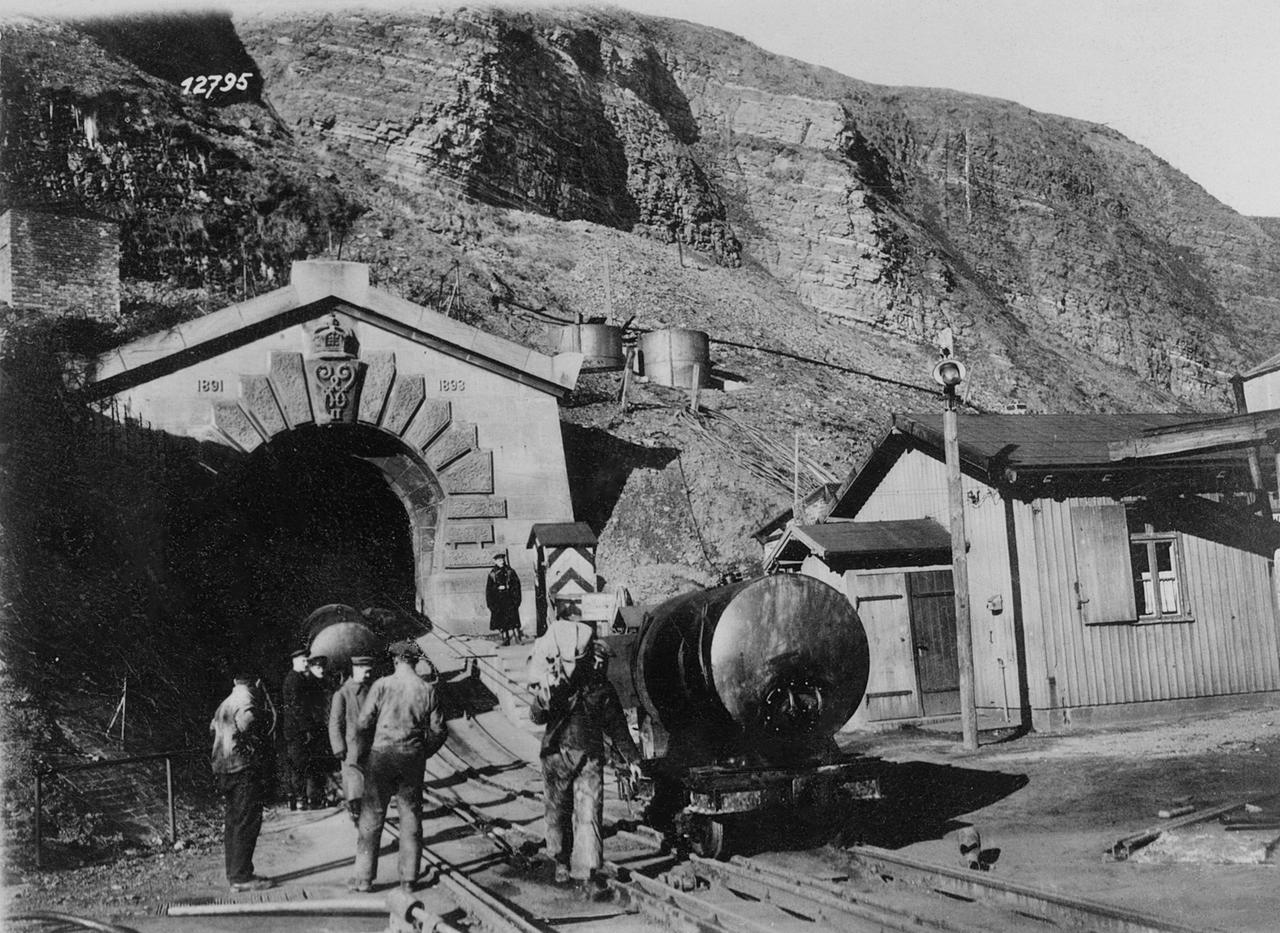 Historisches Schwarzweißfoto eines Tunneleingangs, der in einen großen Fels gehauen ist. In den Eingang verlaufen Schienen, darauf sind eine Lore und mehrere Männer unterwegs.