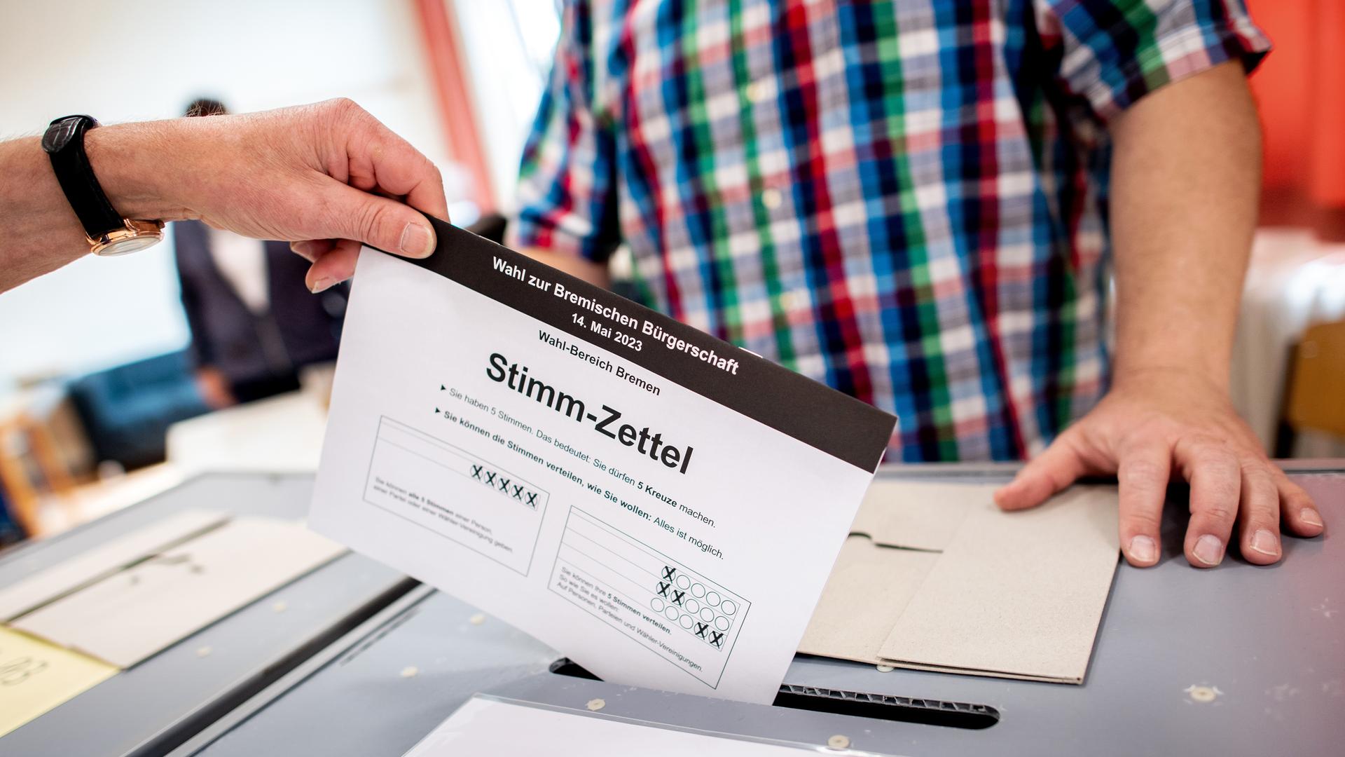 Eine Hand steckt einen Stimmzettel in eine Wahlurne. Im Hintergrund wacht ein Helfer über den korrekten Ablauf. Eine Hand steckt einen Stimmzettel in eine Wahlurne. Im Hintergrund wacht ein Helfer über den korrekten Ablauf.