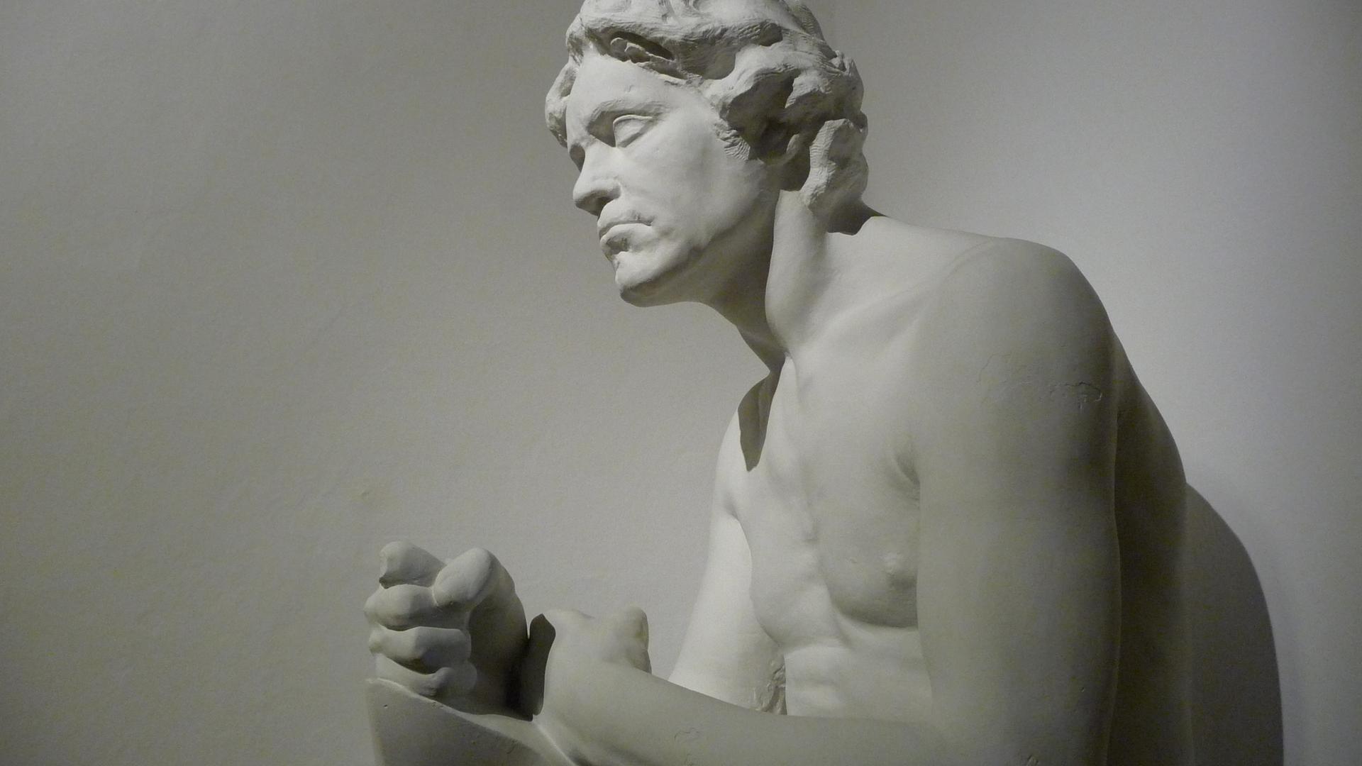 Eine weiße Skulptur, weitlich abgelichtet, von Ludwig van Beethoven als Abguss von Max Klingers Werk aus dem Jahr 1900, wobei er mit freiem Oberkörper und geballter Faust auf einem Stein sitzt und sich leicht nach vorn beugt. Eine weiße Skulptur, weitlich abgelichtet, von Ludwig van Beethoven als Abguss von Max Klingers Werk aus dem Jahr 1900, wobei er mit freiem Oberkörper und geballter Faust auf einem Stein sitzt und sich leicht nach vorn beugt.