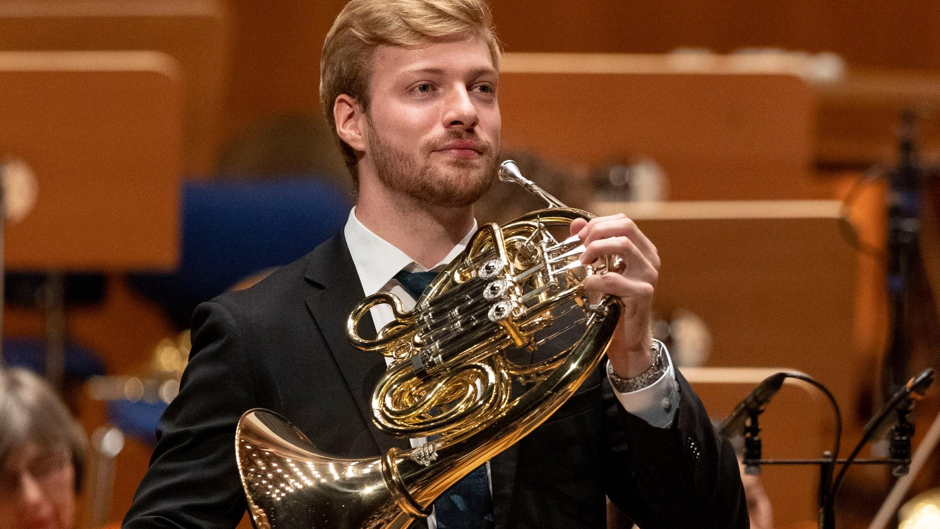 Ein junger Mann mit blonden Haaren steht mit seinem Horn auf der Bühne, kurz bevor er seinen Einsatz hat. Ein junger Mann mit blonden Haaren steht mit seinem Horn auf der Bühne, kurz bevor er seinen Einsatz hat.