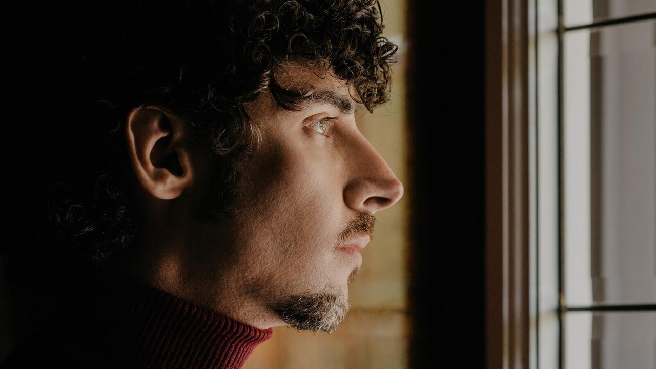 Ein junger Mann mit kurzen schwarzen Locken blickt aus einem Fenster. Wir sehen sein Gesicht im Profil. Ein junger Mann mit kurzen schwarzen Locken blickt aus einem Fenster. Wir sehen sein Gesicht im Profil.