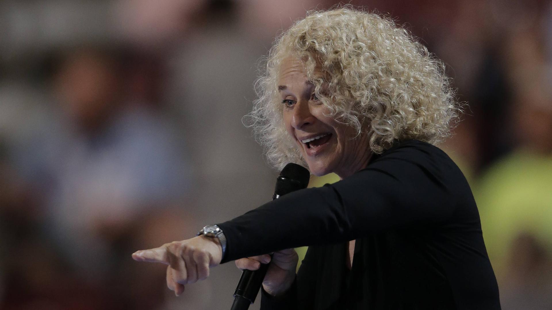 Sängerin Carole King steht auf einer Bühne, hält mit der linken Hand ein Mikrofon und zeigt mit der rechten Rand auf den Bereich vor der Bühne. Sie hat helle, lockige Haare. Sängerin Carole King steht auf einer Bühne, hält mit der linken Hand ein Mikrofon und zeigt mit der rechten Rand auf den Bereich vor der Bühne. Sie hat helle, lockige Haare.