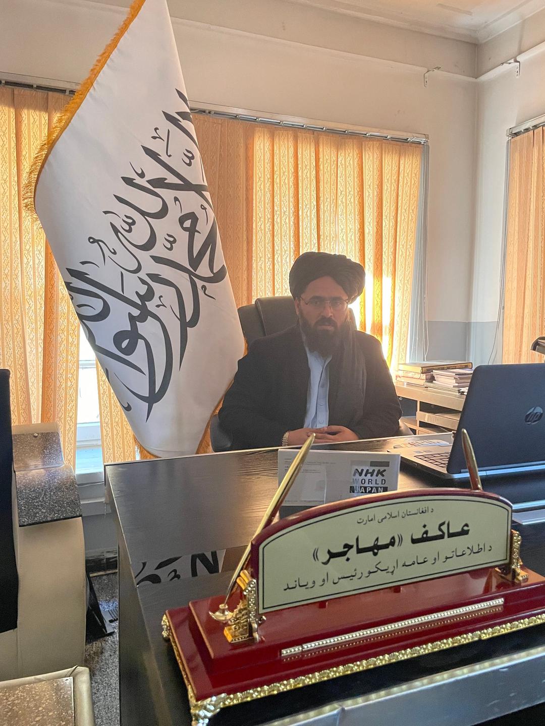 Ein Mann mit Turban und schwarzem Bart sitzt neben einer Fahne in einem Büro.