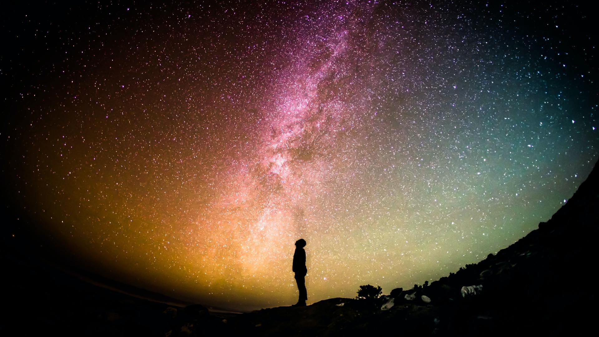 Unter einem bunten Sternenhimmel ist die Silhouette einer Person, die nach oben schaut, zu erkennen. Unter einem bunten Sternenhimmel ist die Silhouette einer Person, die nach oben schaut, zu erkennen.