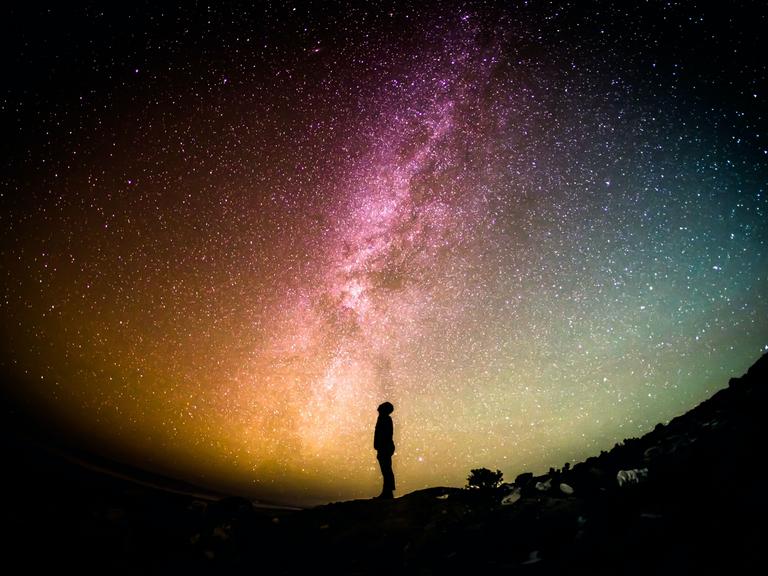 Unter einem bunten Sternenhimmel ist die Silhouette einer Person, die nach oben schaut, zu erkennen.