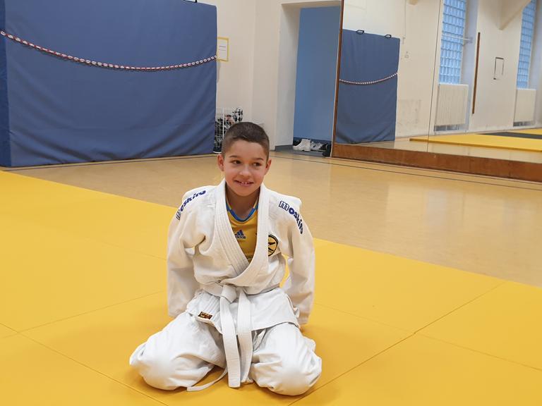 Der elfjährige Pavlo sitzt mit einem Judoanzug, einem Keikogi, bekleidet auf einer Judomatte in einer Turnhalle.