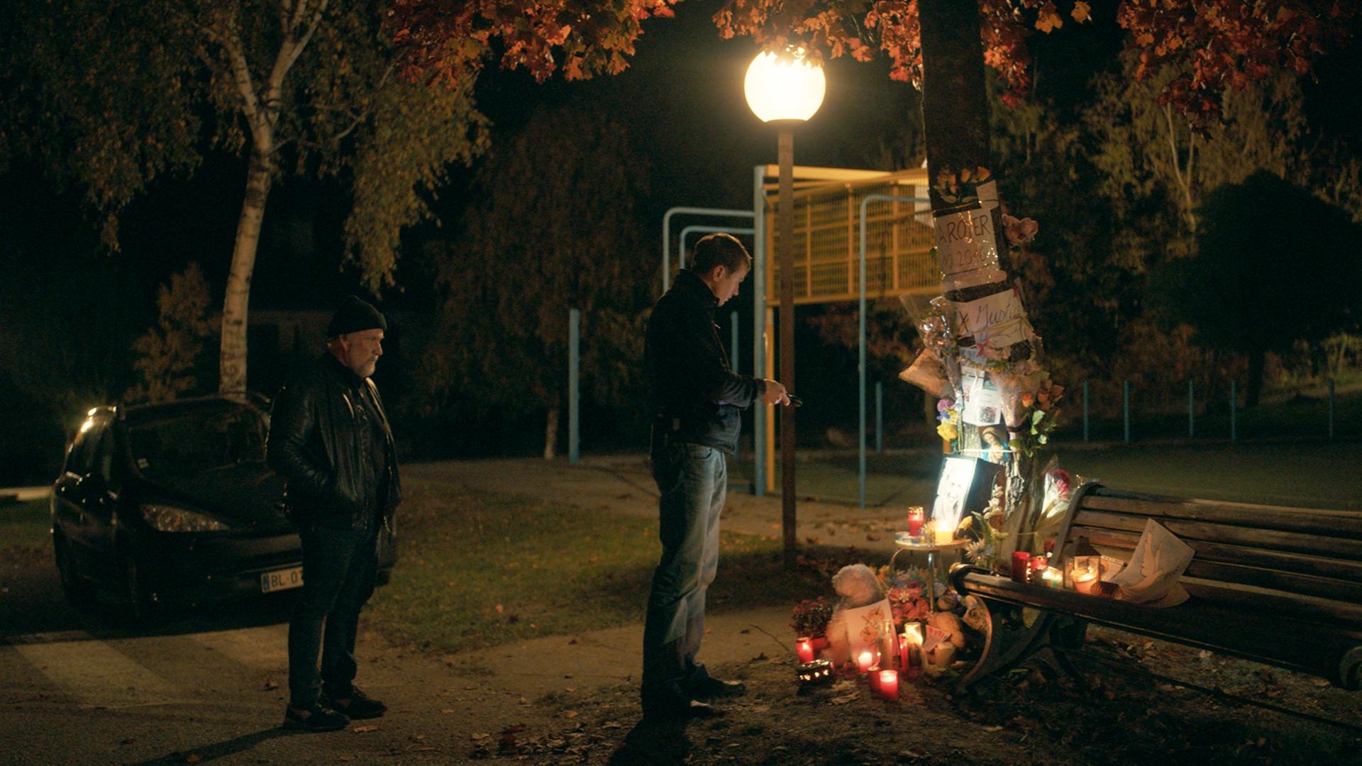 Zwei Männer stehen im Dunkeln am Tatort eines Mordfalls. Dort brennen Kerzen, die an das Opfer erinnern. Zwei Männer stehen im Dunkeln am Tatort eines Mordfalls. Dort brennen Kerzen, die an das Opfer erinnern.
