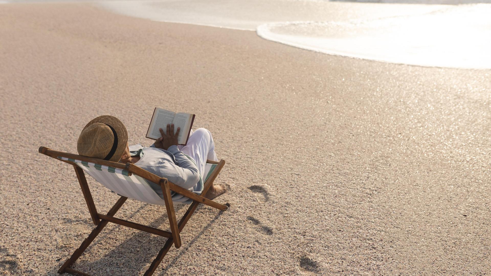 Eine Frau in einem Liegestuhl liest ein Buch am Strand.
