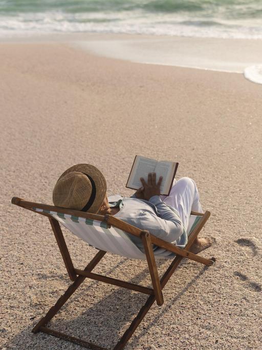 Eine Frau in einem Liegestuhl liest ein Buch am Strand.