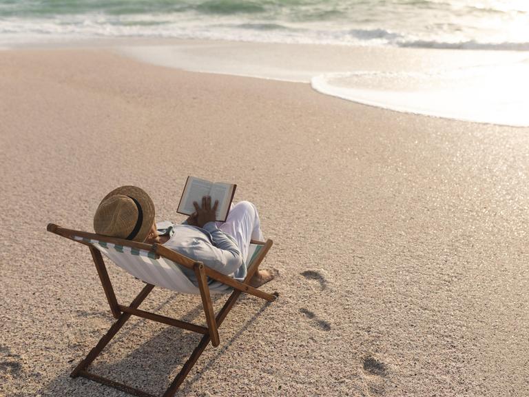 Eine Frau in einem Liegestuhl liest ein Buch am Strand.