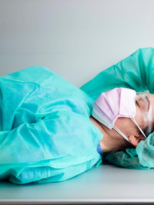 Eine Ärztin liegt in Schutzkleidung erschöpft auf einer Metallbank.