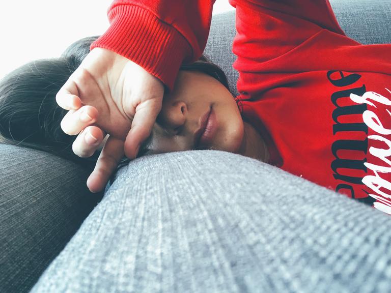 Eine Frau mit rotem Sweater liegt auf dem Sofa und hält sich die Hand vor die Augen.