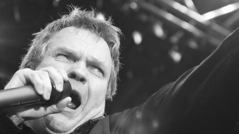 Der Sänger Meat Loaf in Nahaufnahme. Er ruft ins Mikrofon, das er mit der linken Hand dicht an den Mund hält. Mit dem rechten Zeigefinger zeigt er ins Publikum.