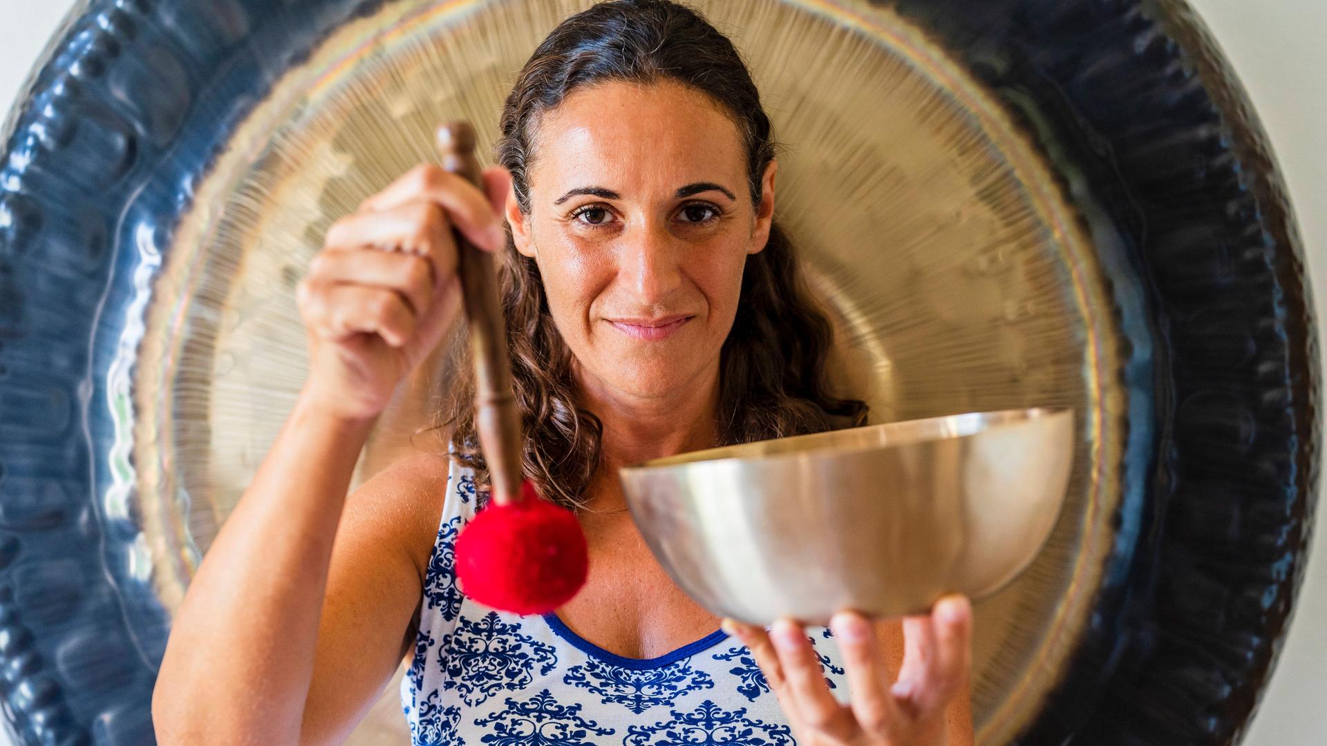 Eine junge Frau hält vor einem großen Gong eine Klangschale lächelnd in die Kamera, die sie gerade mit einem Schläger zum Klingen gebracht hat. Eine junge Frau hält vor einem großen Gong eine Klangschale lächelnd in die Kamera, die sie gerade mit einem Schläger zum Klingen gebracht hat.