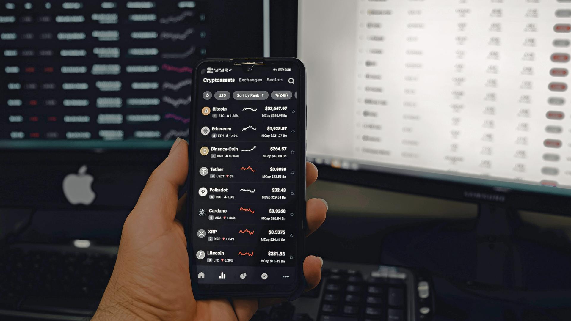 Eine Hand hält ein Smartphone, auf dessen Display mehrere Kryptowährungskurse zu sehen sind, im Hintergrund sind zwei Computer moniture zu sehen.
Eine Hand hält ein Smartphone, auf dessen Display mehrere Kryptowährungskurse zu sehen sind, im Hintergrund sind zwei Computer moniture zu sehen.